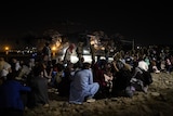 A group of Afghan evacuees sit on the ground with a large plane in the background. 