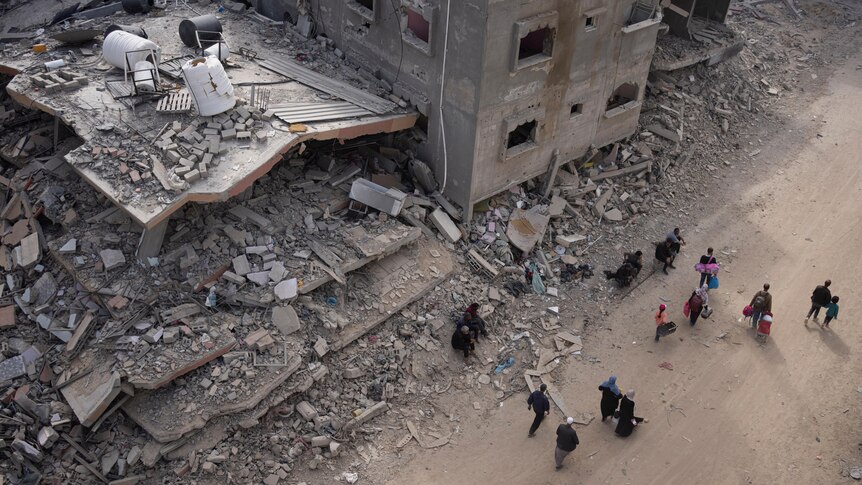 Above-the-head shot of a dozen people walking down a street next to bombed buildings turned to rubble