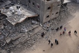 Above-the-head shot of a dozen people walking down a street next to bombed buildings turned to rubble
