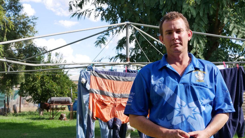 A man stands in front of his clothes line in the backyard wearing a mining union blue shirt, looks serious.