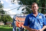A man stands in front of his clothes line in the backyard wearing a mining union blue shirt, looks serious.