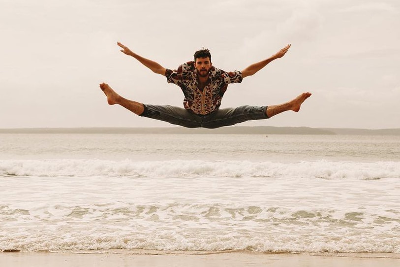 a young man jumps with his legs in splits and arms outstretched at the beach.