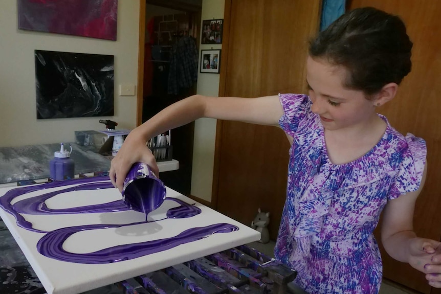 Young girl smiling as she pours purple paint from a cup onto a canvas
