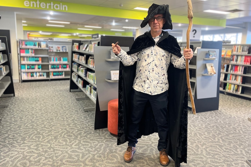 A man in a library dressed in a wizard's cape and hat, holding a wooden staff.