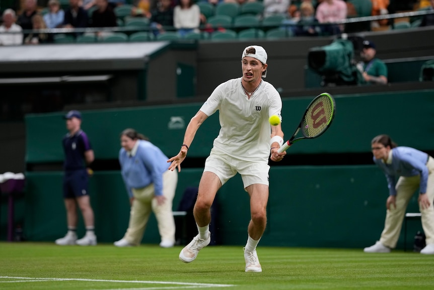 A cap-wearing tennis player blocks a left-handed forehand return from behind the baseline as he plays at Wimbledon.
