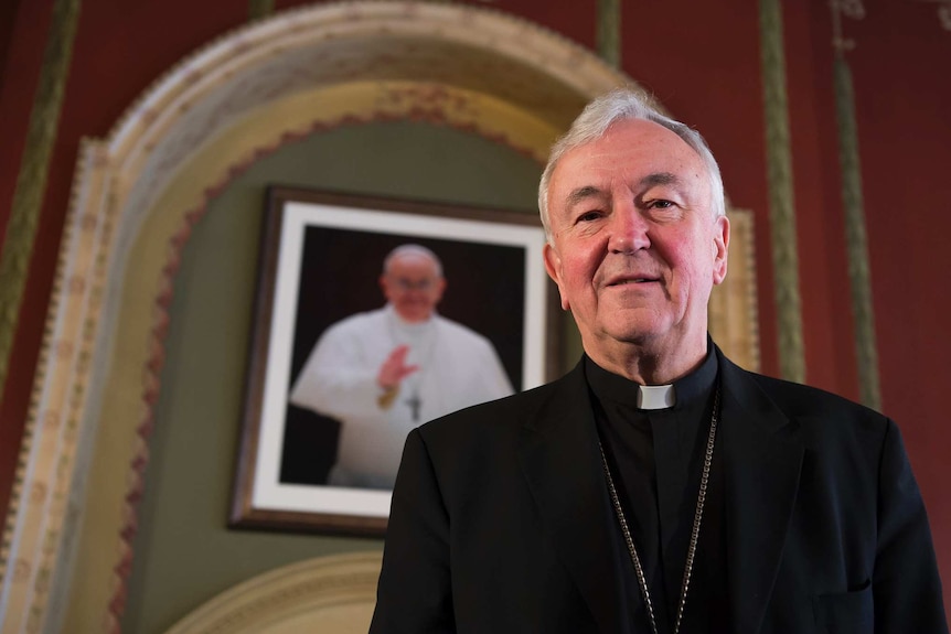 You view Vincent Nichols in a black suit with a priest's white collar standing in front of a portrait of Pope Francis.