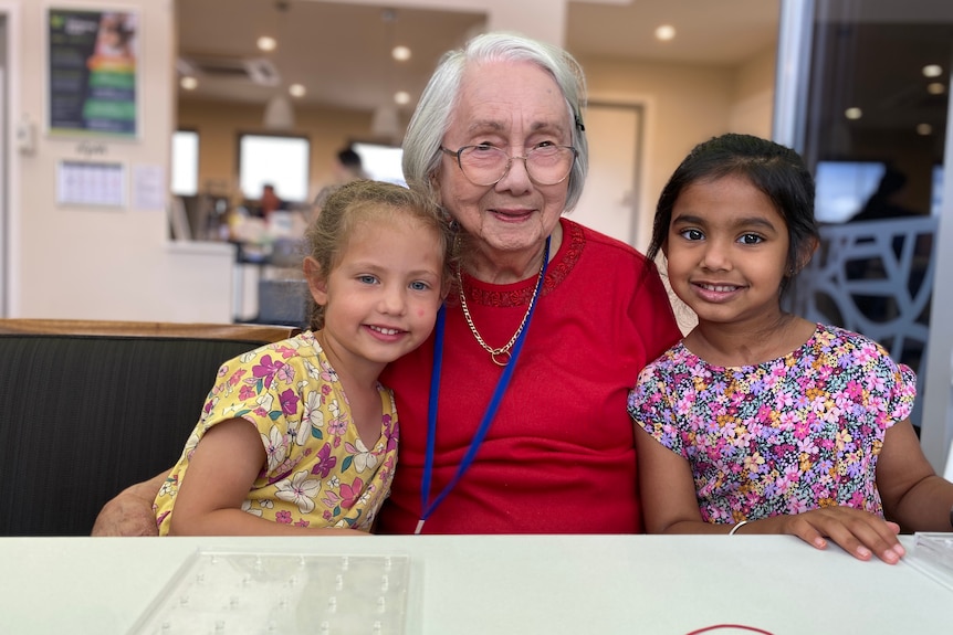 An image of Esther Baulch, 89, with a white bob, wearing a red top, sitting next to two children in colourful floral shirts
