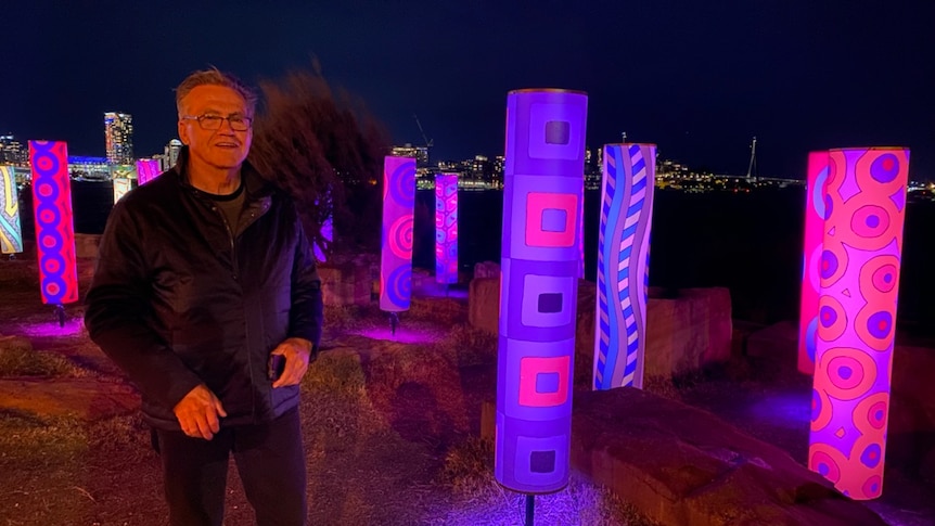 Warwick Keen stands alongside pink and blue light up cylinders with his art on them at night.