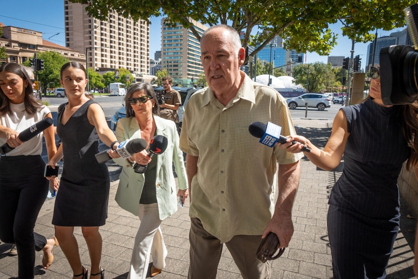A man with a yellow checkered shirt walks outside of court surrounded by female journalists holding microphones.