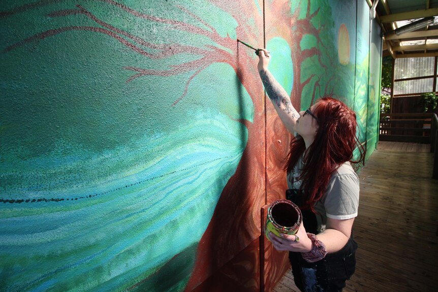 a woman with long red hair holds a jar of paint as she paints a picture of a tress on a wall