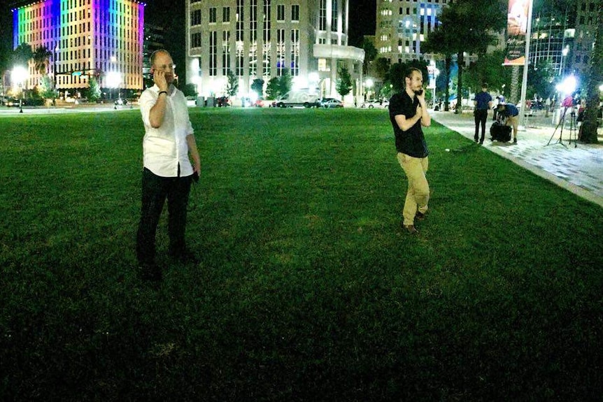 Two reporters standing on grass outside nightclub on phones.