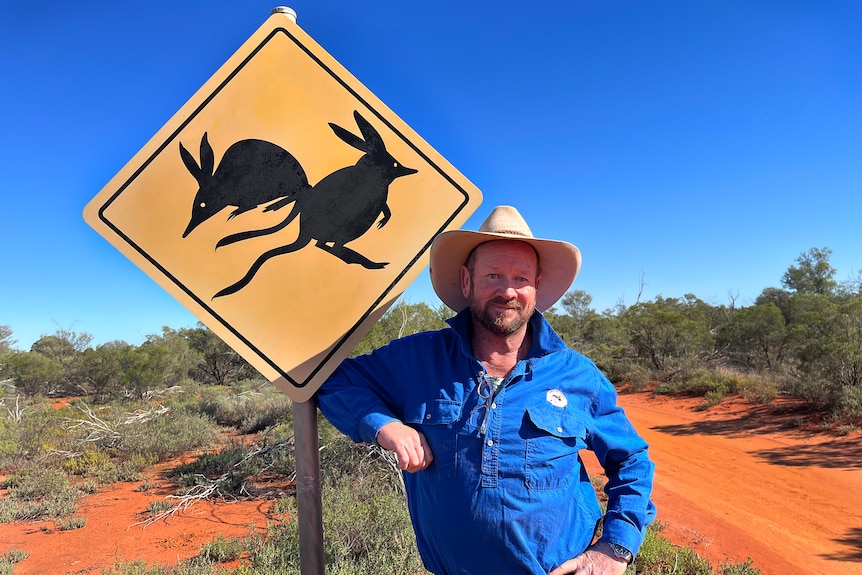 A man in a blue shirt leaning against a yellow sign with bilbies on it