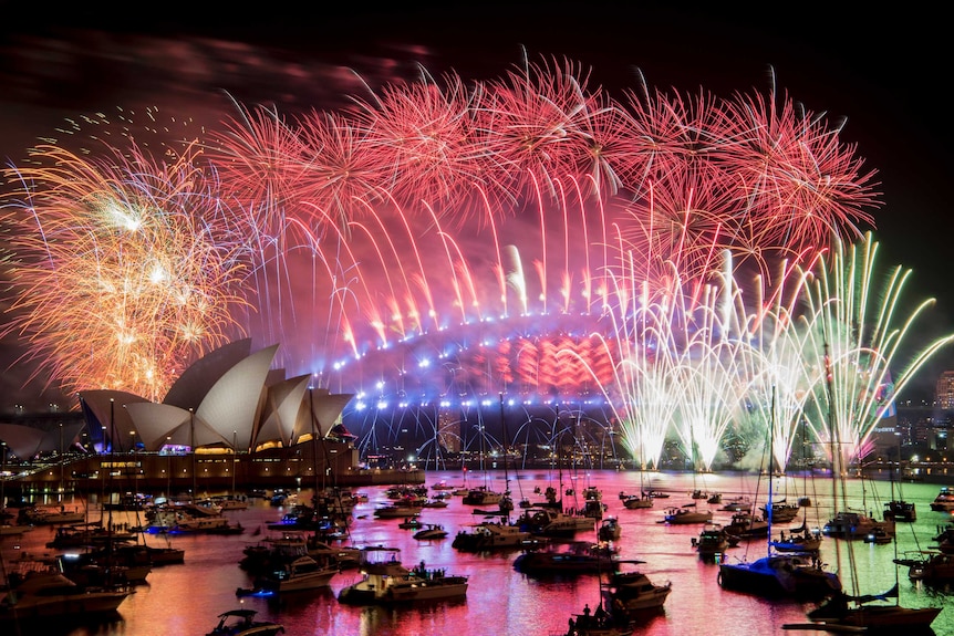 Red fireworks explode from the Sydney Harbour Bridge, with orange-yellow fireworks going off over Opera House and green on right