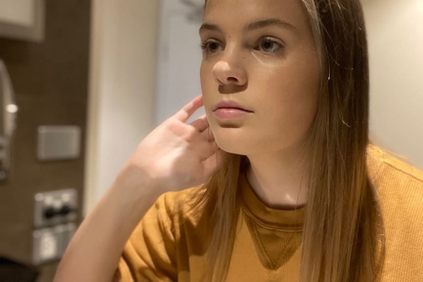 A teenager girl looks into an unseen mirror. She has one hand in her hair and a vacant expression on her face.