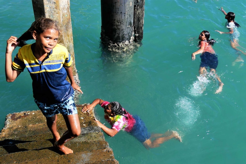 A girl looks up on the steps of the jetty while three girls swim below