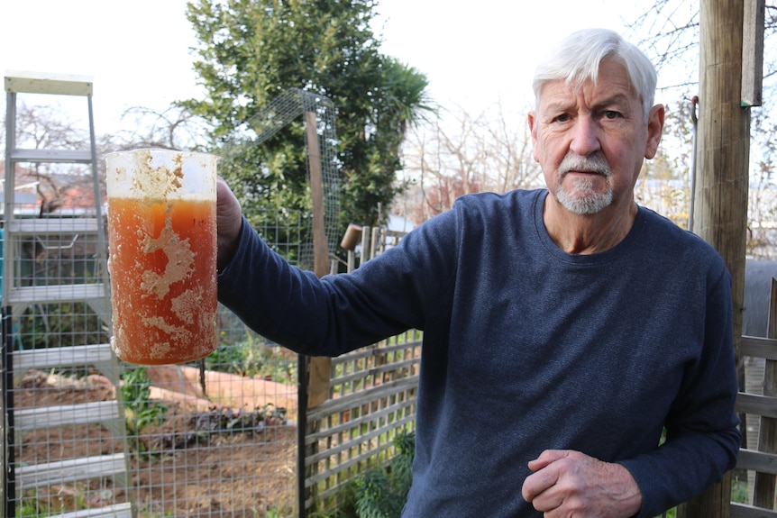 Gerry holds a jug of amber liquid up, in his backyard.