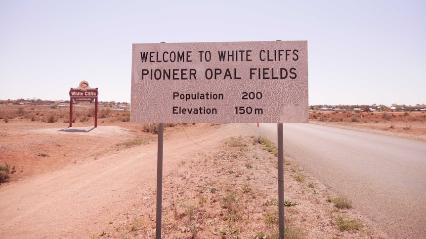 A sign reading 'Welcome to White Cliffs - Pioneer Opal Fields' by the side of a road.