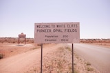 A sign reading 'Welcome to White Cliffs - Pioneer Opal Fields' by the side of a road.