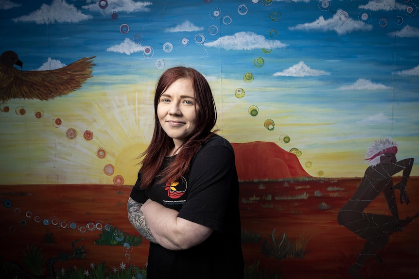 A woman smiling in front of a mural she has painted