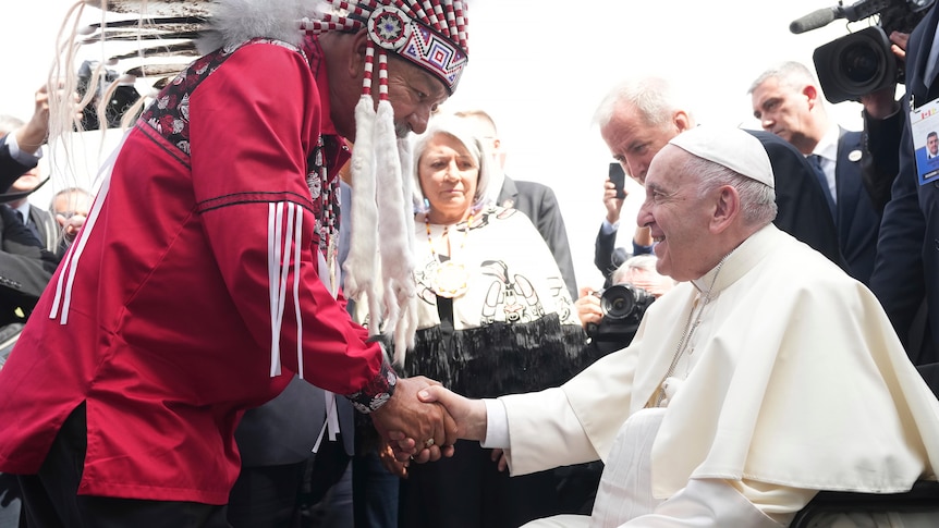 Dressed in robes and in a wheelchair Pope Francis shakes hands with a man in red traditional dress. 