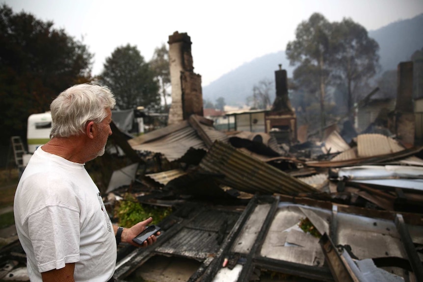A man stands in a white shirt with his head turned towards the rubble of a house that was destroyed in a bushfire.