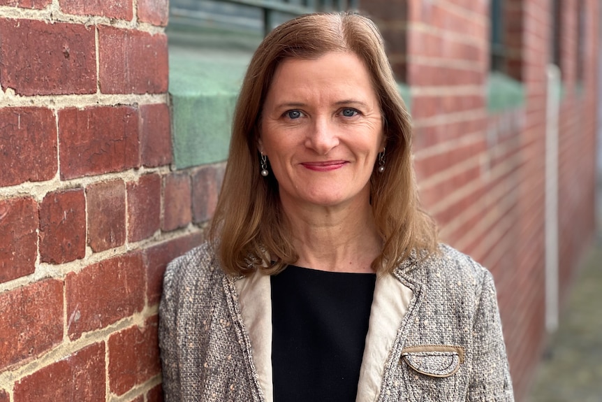 Woman wearing a black top and light grey blazer leaning against a brick wall.