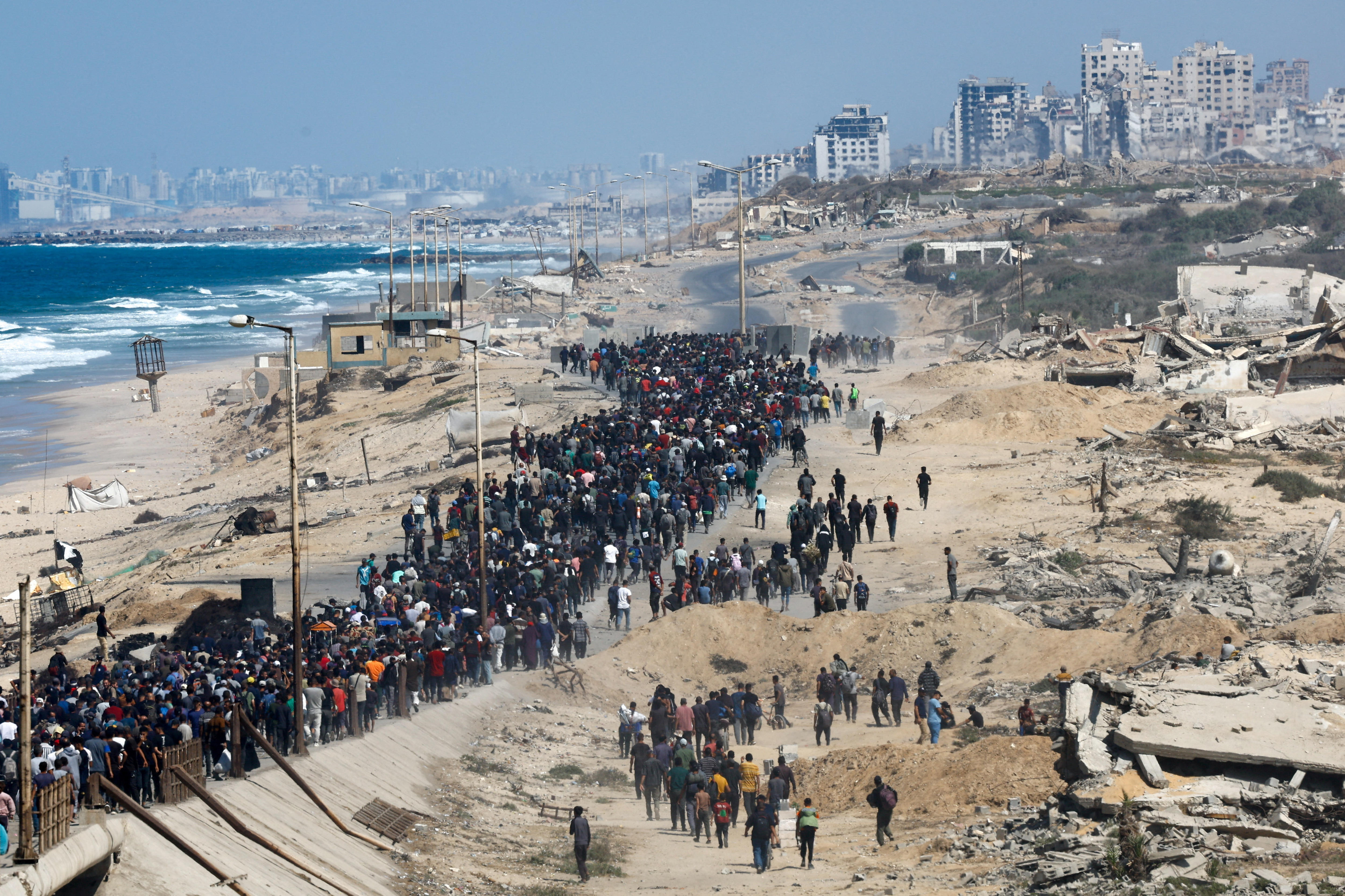 Hundreds of Palestinians walk along a coastal road, beside war ruins