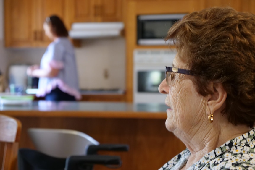  Bell Cares client Joan Hunter sitting in her dining room while her carer Sarah Heathwood washes the dishes