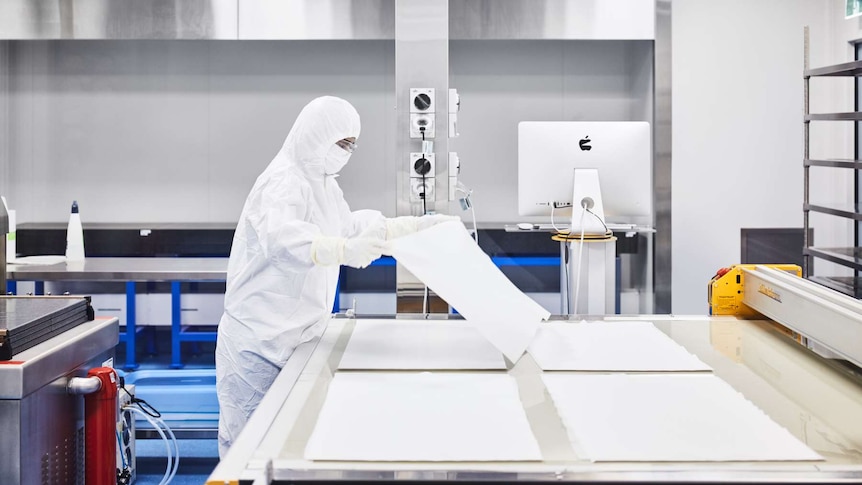 A lab technician dressed in white scrubs holds up a white sheet of material in a laboratory.