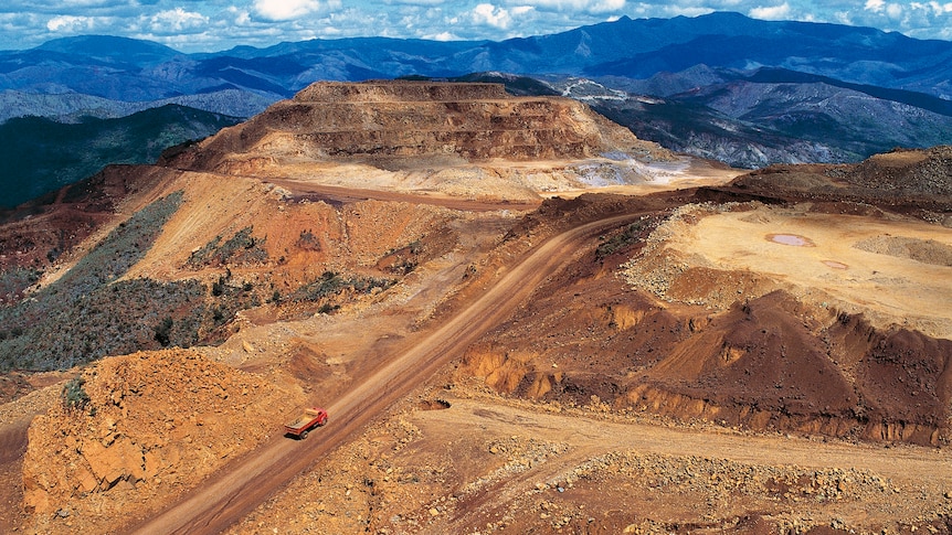 Aerial photo showing a truck dwarfed by the enormous mined land around it. In the distance are blue-green hills with vegetation.