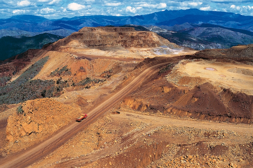Aerial photo showing a truck dwarfed by the enormous mined land around it. In the distance are blue-green hills with vegetation.