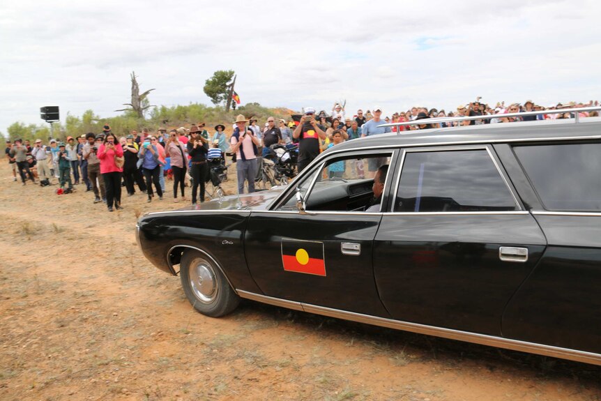 A black hearse, with an Aboriginal flag painted on its side, drives while a crowd watches on.