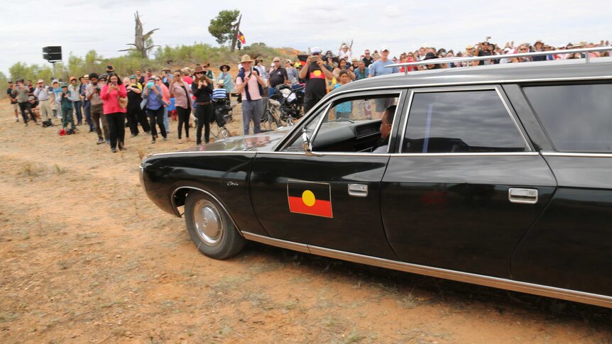 A black hearse, with an Aboriginal flag painted on its side, drives while a crowd watches on.