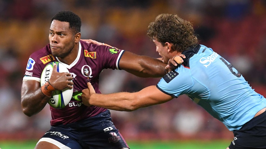 Filipo Daugunu holds a rugby ball under one arm and holds a player in a blue shirt with the other