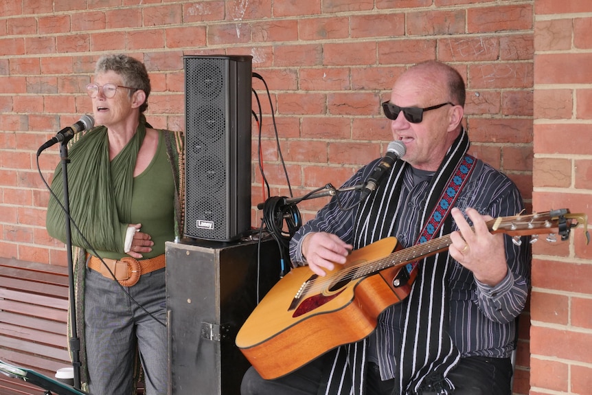 A man sititng down playing guitar and a woman singing out the front of a brick building 