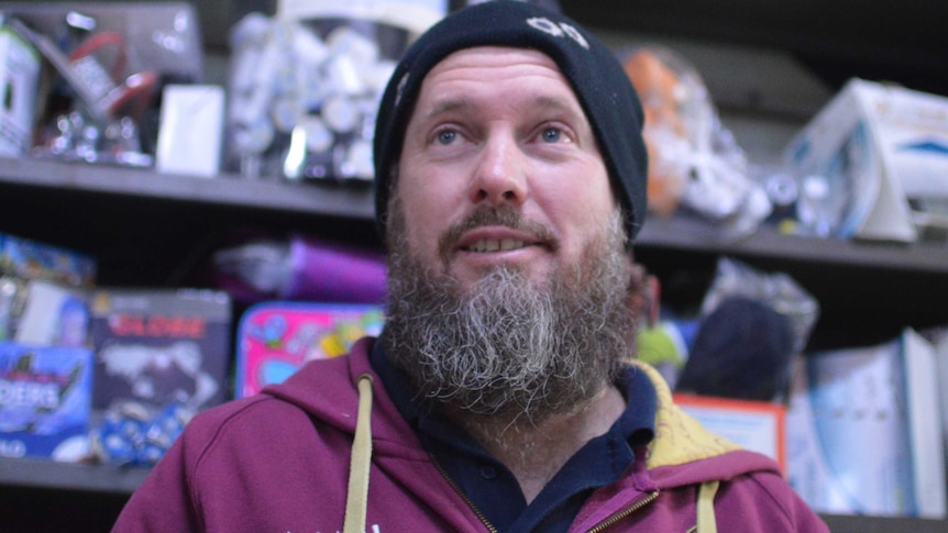 A man with a beard, wearing a beanie and a maroon hoodie, standing in front of a shop shelf.