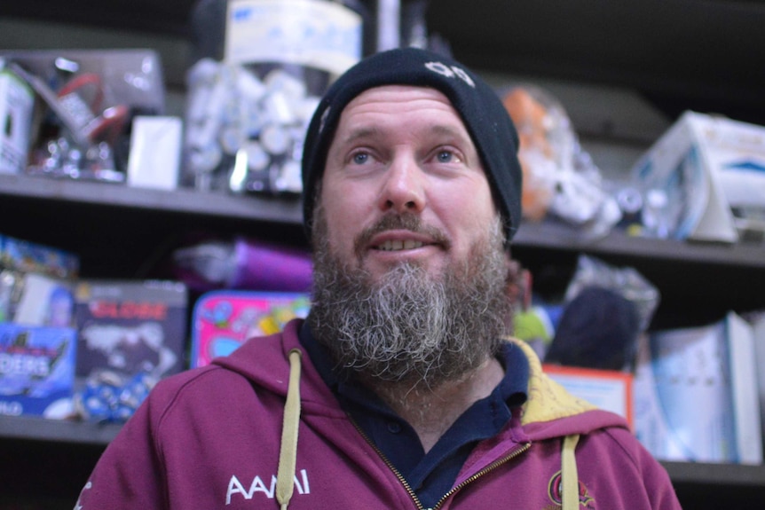 A man with a beard, wearing a beanie and a maroon hoodie, standing in front of a shop shelf.