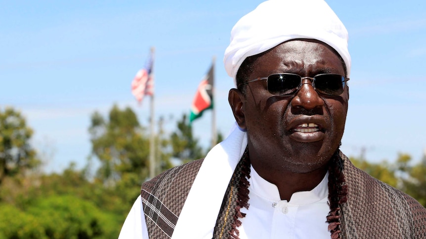 Malik Obama, Barack Obama's half-brother, stands in front of Kenyan and American flags on a sunny day.