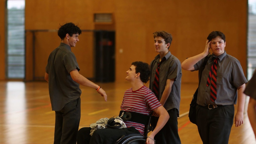 Boy in a wheelchair wearing a stripy shirt laughs with three friends in school uniform.