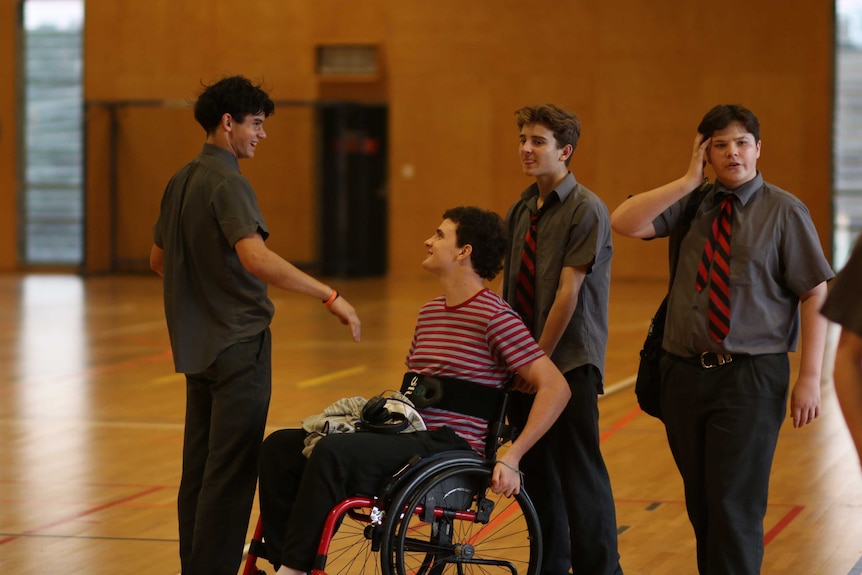 Boy in a wheelchair wearing a stripy shirt laughs with three friends in school uniform.