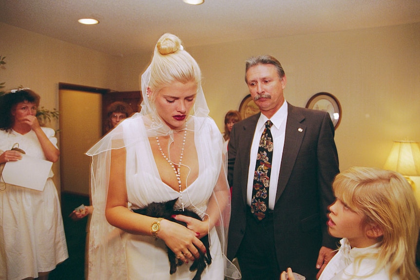A young woman with blonde hair wearing a white low-cut dress attends a funeral for her late husband. She is holding a small dog.