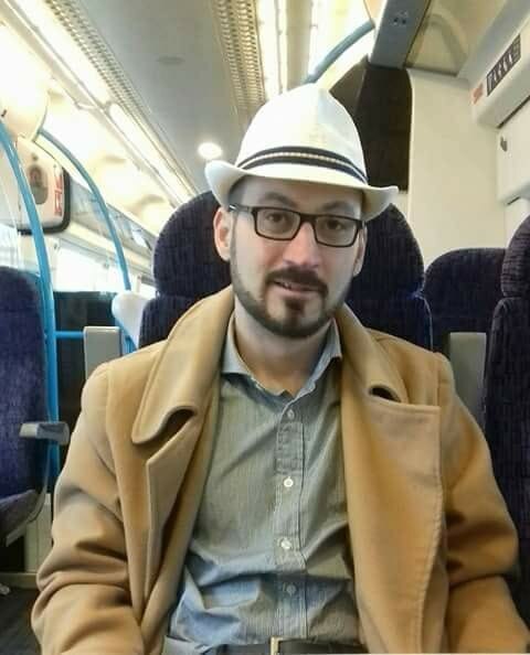 Un hombre barbudo con gafas lleva un sombrero de fieltro mientras está sentado en un tren.