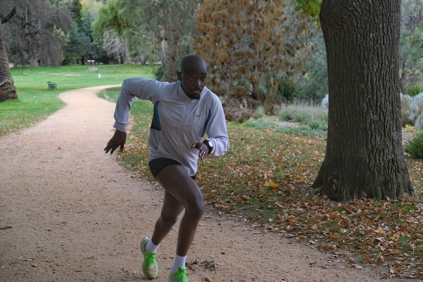 A man wearing a white top, black shorts and green shoes runs along a path in a park.