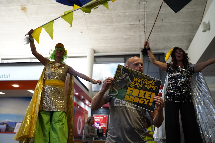 Two performers on stilts wear sparkling costumes in the colours of Australia and New Zealand, while a man shows an ad.