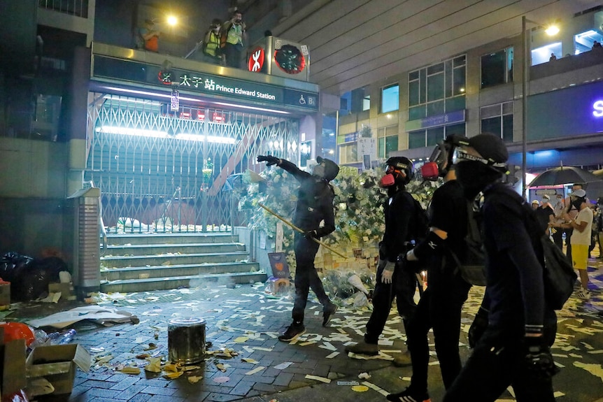 A shuttered metro station entrance shows four Hong Kong protesters in black throwing a projectile to a police station.
