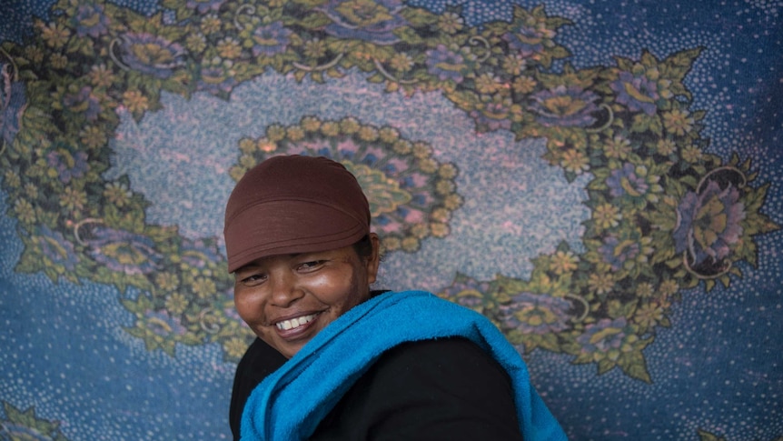 A close up of Aina wearing a brown cap against a hanging blue sheet with a floral pattern on it.
