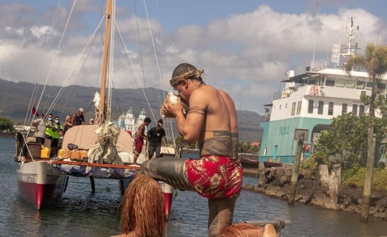 Man kneels infrong of sea canal dressed in ie lavalava, wearing the pe'a, a Samoan tatoo from his waist to knees, holds shell. 