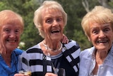 Three sisters smile happily at the camera as they hold glasses of champagne.