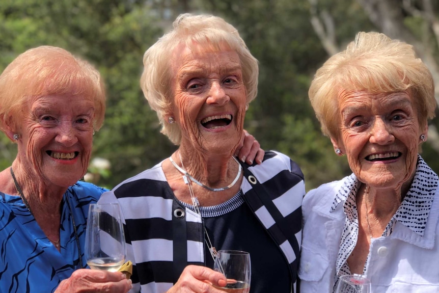 Three sisters smile happily at the camera as they hold glasses of champagne.