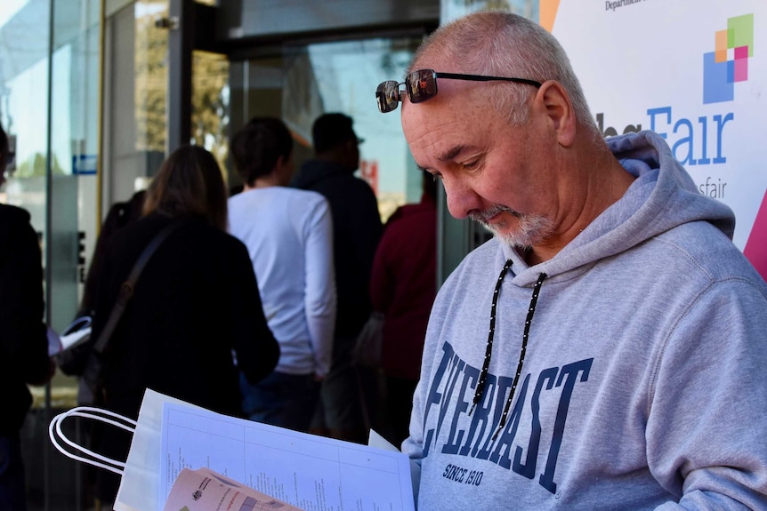 A man looks at job advertisements on a sheet in his hands, while a line of people enter the Jobs Fair.